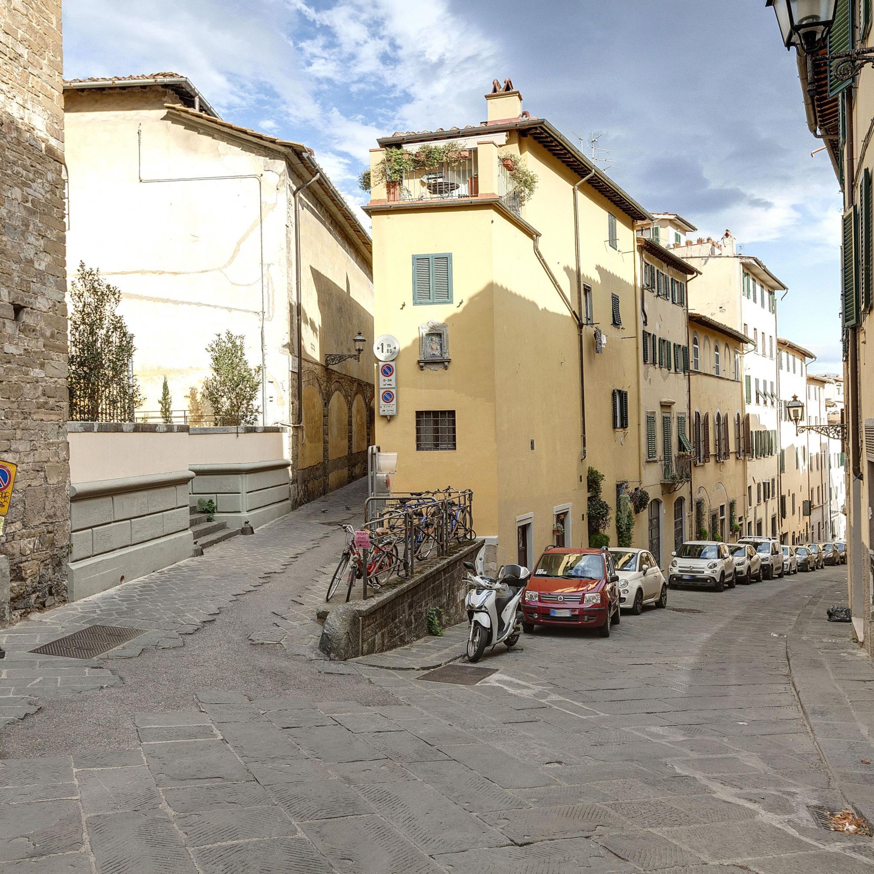 A narrow street lined with historic buildings, a parked scooter, and several cars.