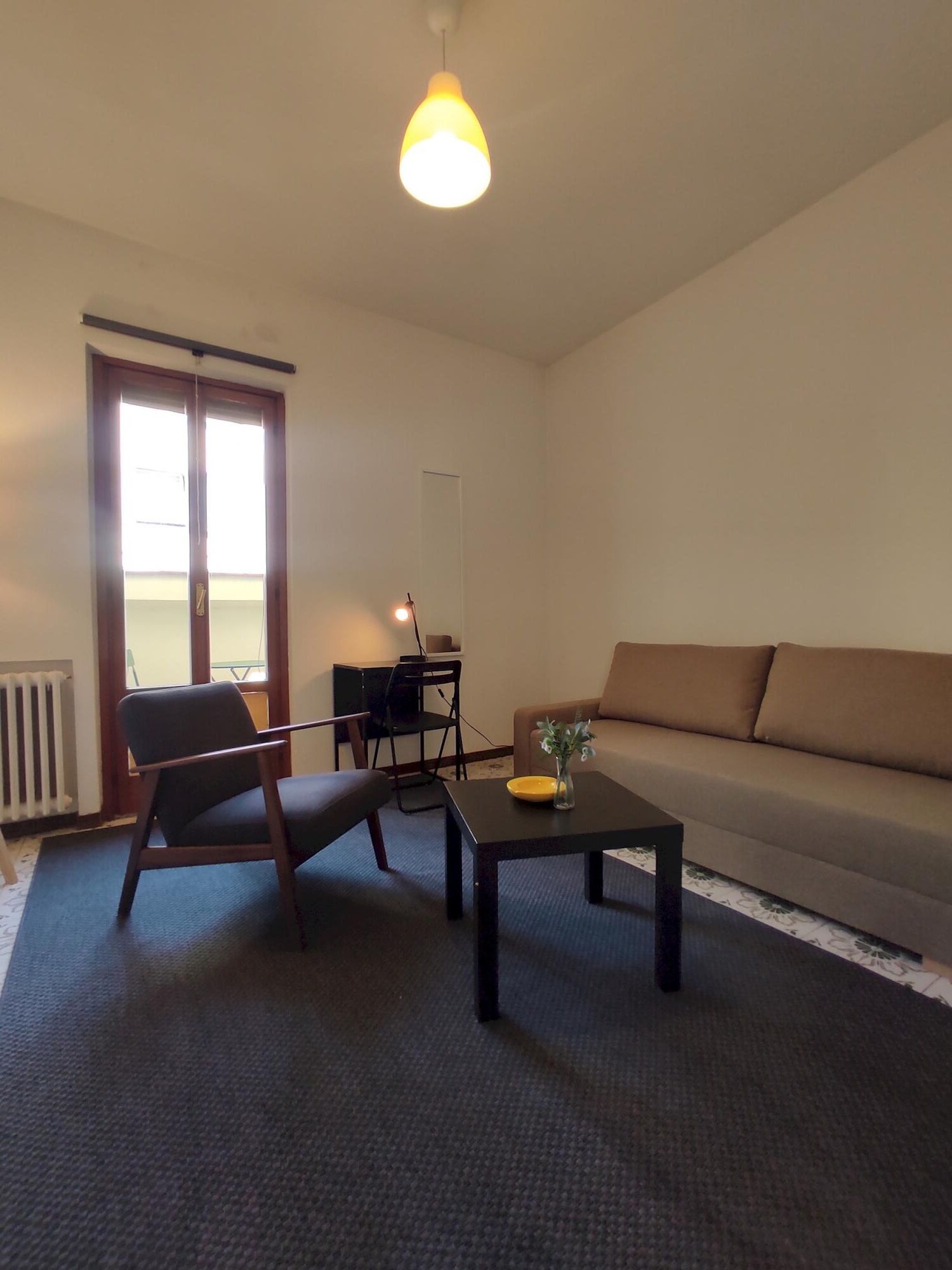 A living room with a sofa, a chair, a coffee table, and a lamp. There is a door leading to a balcony.