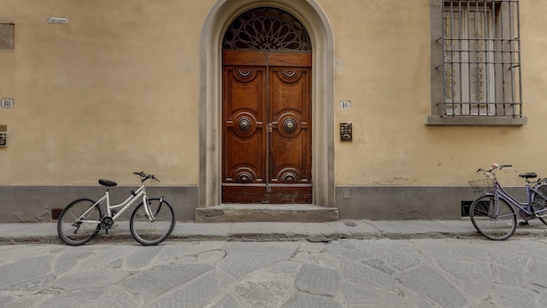 A building with a large wooden double door, two bicycles parked in front, and a window with metal bars. - Greci 16 III in Firenze (Florence)