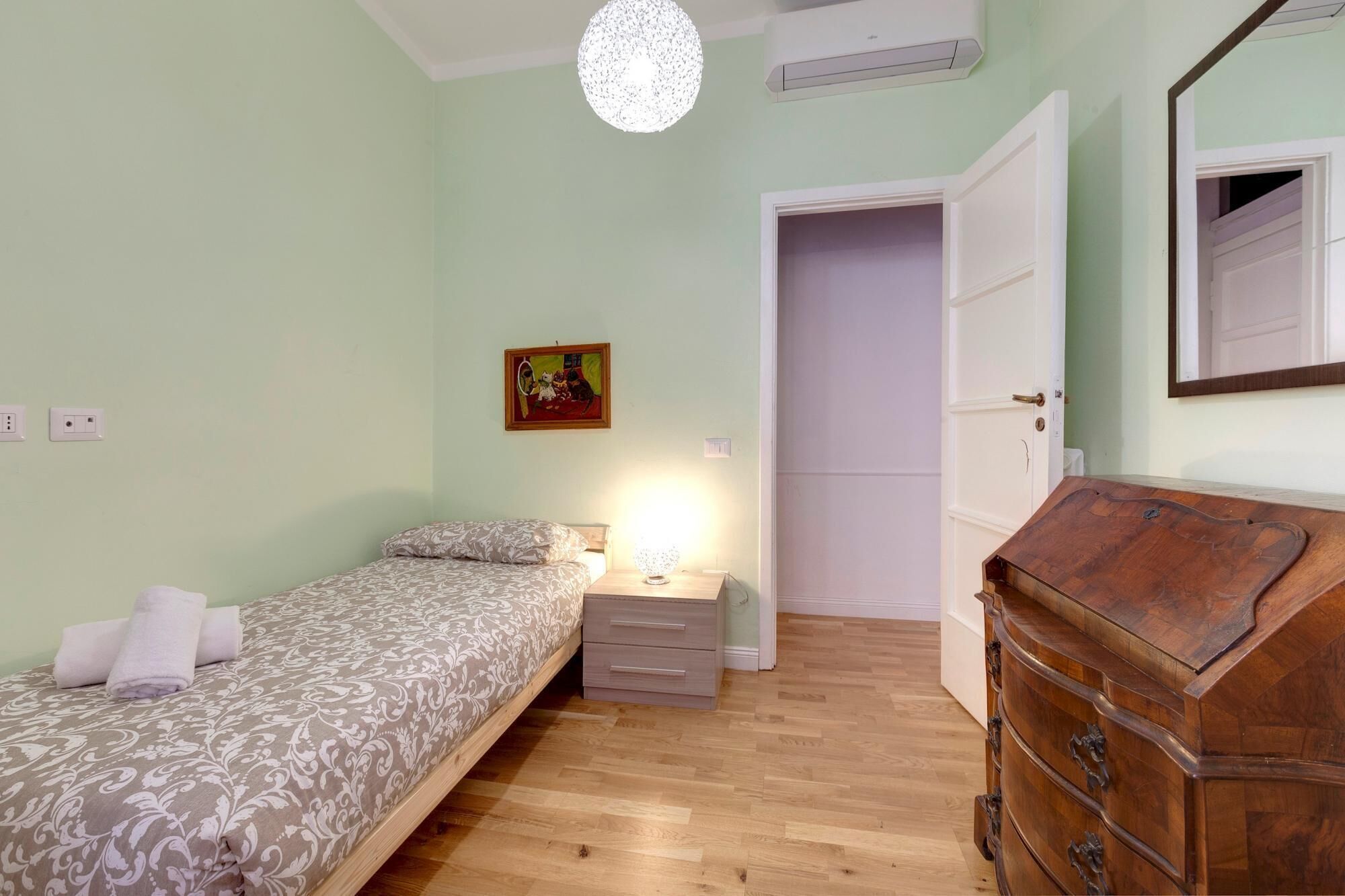 A bedroom with a bed, bedside table, lamp, dresser, and a door leading to another room.