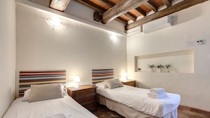 Une chambre d’hôtel avec deux lits, chacun muni d’un oreiller et d’une serviette pliée. On y trouve des tables de chevet en bois avec des lampes, une tête de lit à rayures et une petite étagère avec des plantes.