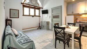 Un espacio habitable compacto con comedor, sofá, televisión y cocina, que incluye un reloj y una ventana.
