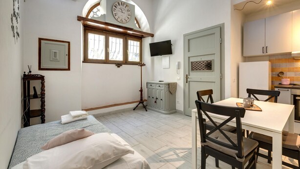Una habitación con cama, una mesa con sillas, un reloj, un televisor y un ventanal con cortinas.