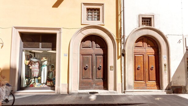 A street view with two arched wooden doors, numbered 54 and 52, and a shop with mannequins and bags.