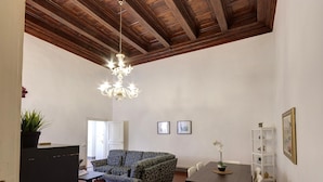 A living room with a wooden ceiling, a chandelier, a sofa, a dining table with chairs, and a bookshelf.