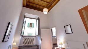 A hotel room with a wooden ceiling, a large window with shutters, a radiator, a bed with a nightstand, and a lamp.