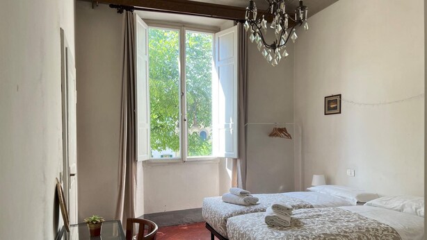 Quarto de hotel com duas camas, um candelabro, uma janela com cortinas, uma mesinha com uma planta e uma cadeira.