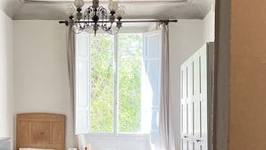 Quarto com mesa de madeira, cadeiras, um lustre, janela com cortinas e cama com cabeceira.