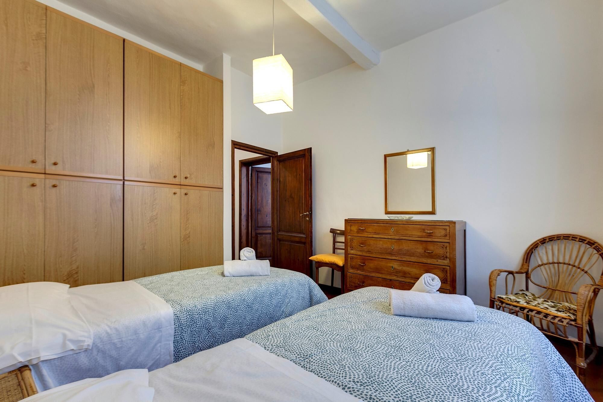 Un dormitorio con dos camas, un armario de madera, un tocador, un espejo y una silla de mimbre.