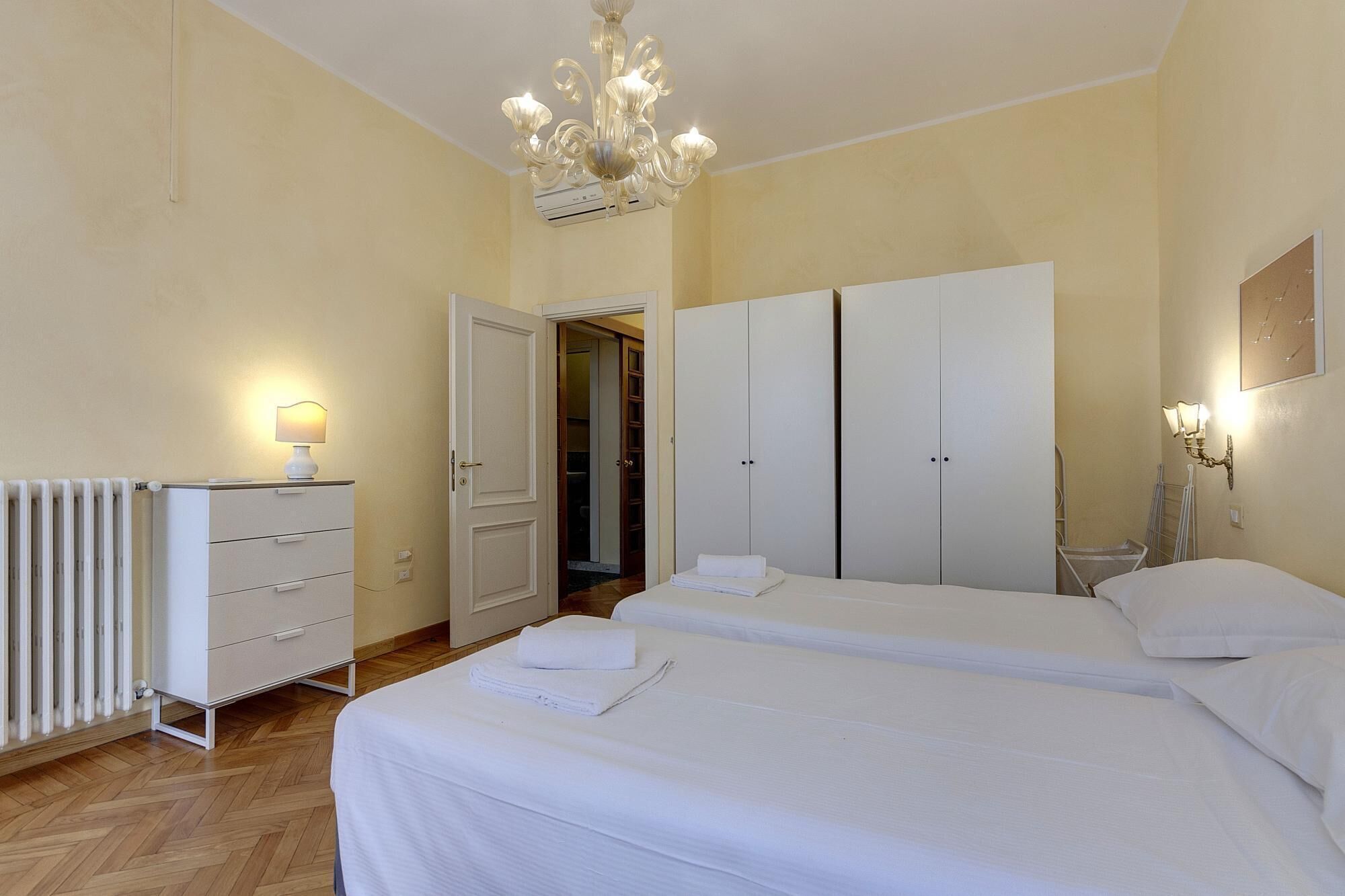 A hotel room with two beds, a radiator, a wardrobe, and a chandelier.