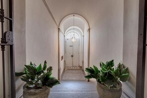 Um corredor com uma porta no final, dois vasos de plantas de cada lado e piso coberto por carpete.