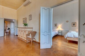 Ett hotellrum med ett trägolv i fiskbensmönster, en säng med vita sängkläder, en flätad fåtölj, ett litet bord med en lampa och ett skåp med dekorativa föremål.