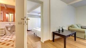 Ett hotellrum med trägolv, en vit säng, ett litet bord med blommor, en grön soffa och ett badrum med handfat och spegel.