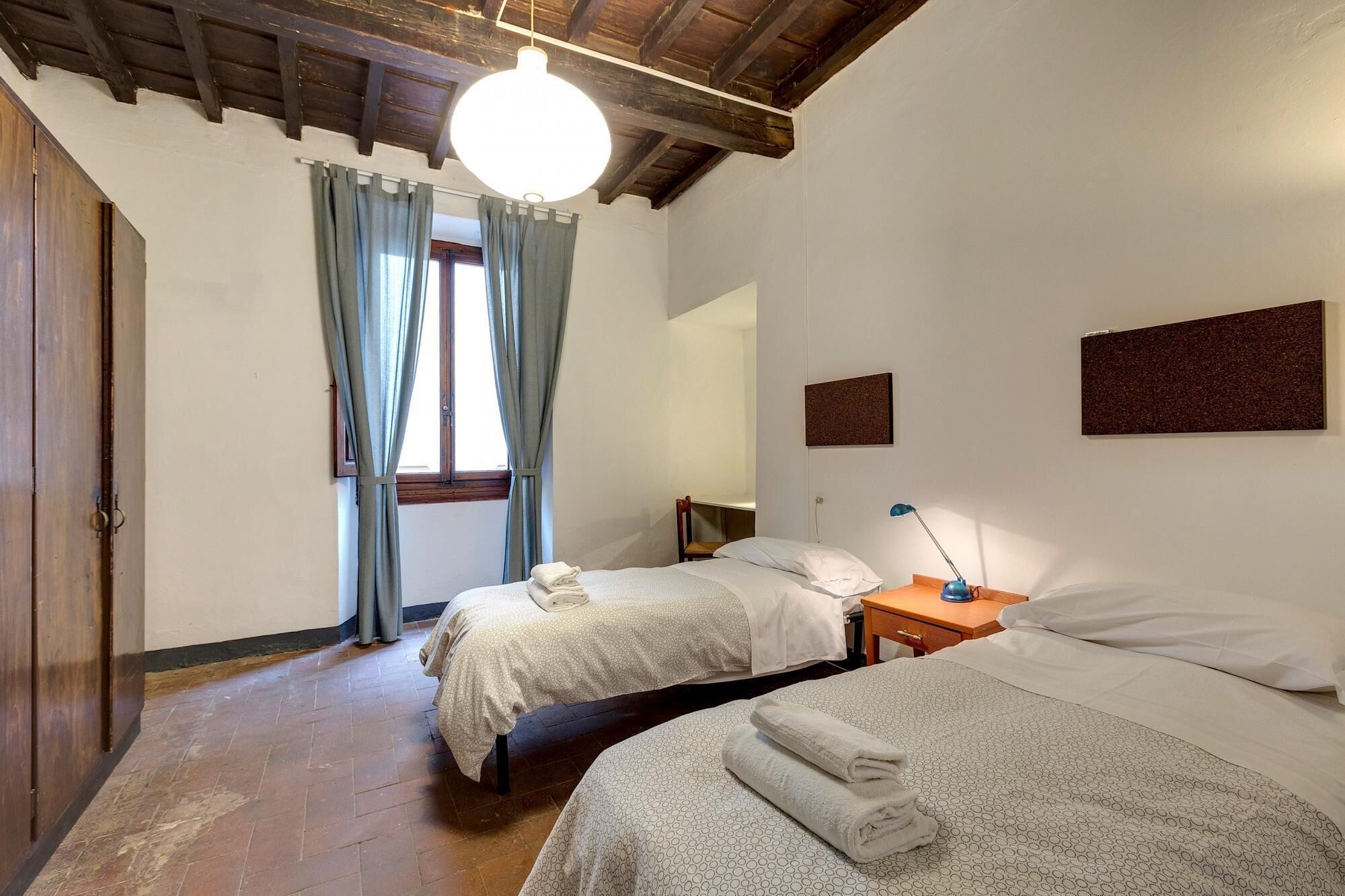 A room with two beds, a wooden door, a window with curtains, a hanging light fixture, and a small bedside table with a lamp.