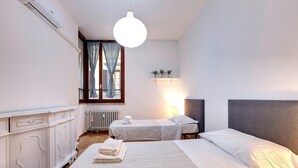 A hotel room with two beds, a radiator, a window with curtains, a hanging light fixture, and a small shelf with plants. - Malenchini 6 in Firenze (Florence)