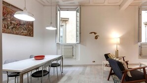 A dining area with a white table, chairs, and a red bowl. There is a painting on the wall, a window with a view, and a lamp.
