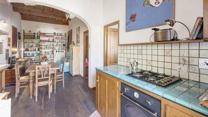 Una cocina con estufa, pileta y salpicadero azulejado. Se observa una estantería llena de libros y un comedor con mesa y sillas.