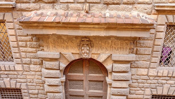 Un bâtiment en pierre doté d’une grande porte en bois, d’une arche en pierre décorative au-dessus de la porte et d’un toit recouvert de tuiles.