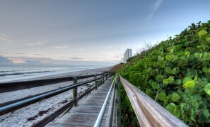 A wooden boardwalk leading to a beach with a metal railing and dense green foliage on one side. - OCEAN VIEW, Swimming Pool & Jacuzzi. 4 bdr, 3 bath house. 3 min walk to beach. (Melbourne Beach)