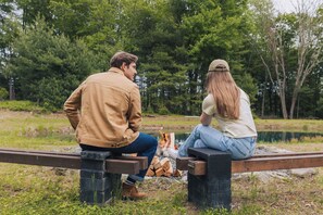 Due persone sedute su una panchina all'aperto, con un fuoco e alberi sullo sfondo.