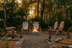 Een boslandschap met een vuurplaats omringd door Adirondack-stoelen.