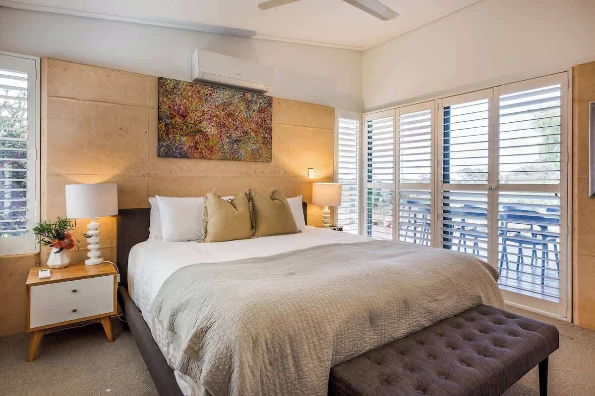 Une chambre à coucher avec un grand lit, deux lampes de chevet, une fenêtre avec des stores et un ventilateur de plafond.