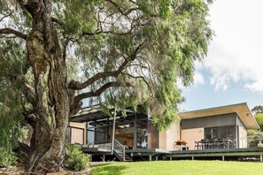 A modern house with a large tree in the foreground, a covered outdoor area with seating, and a clear sky.