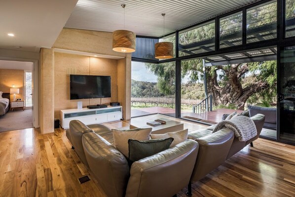 A modern living room with a large flat-screen TV, leather sofas, and a glass-enclosed patio with a view of trees.