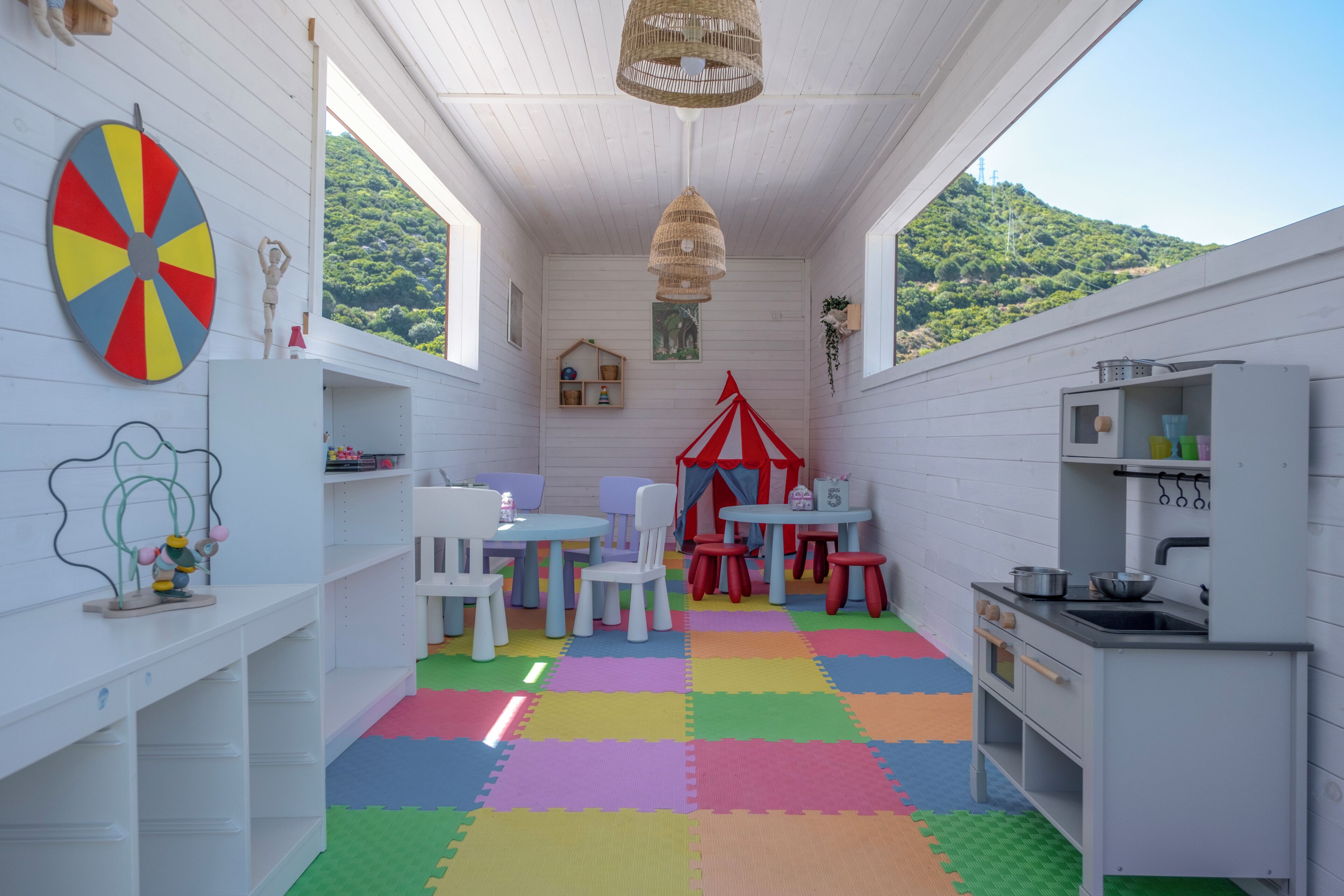 A children's playroom with a colorful floor, white furniture, a small kitchen area, and a view of a green hill.