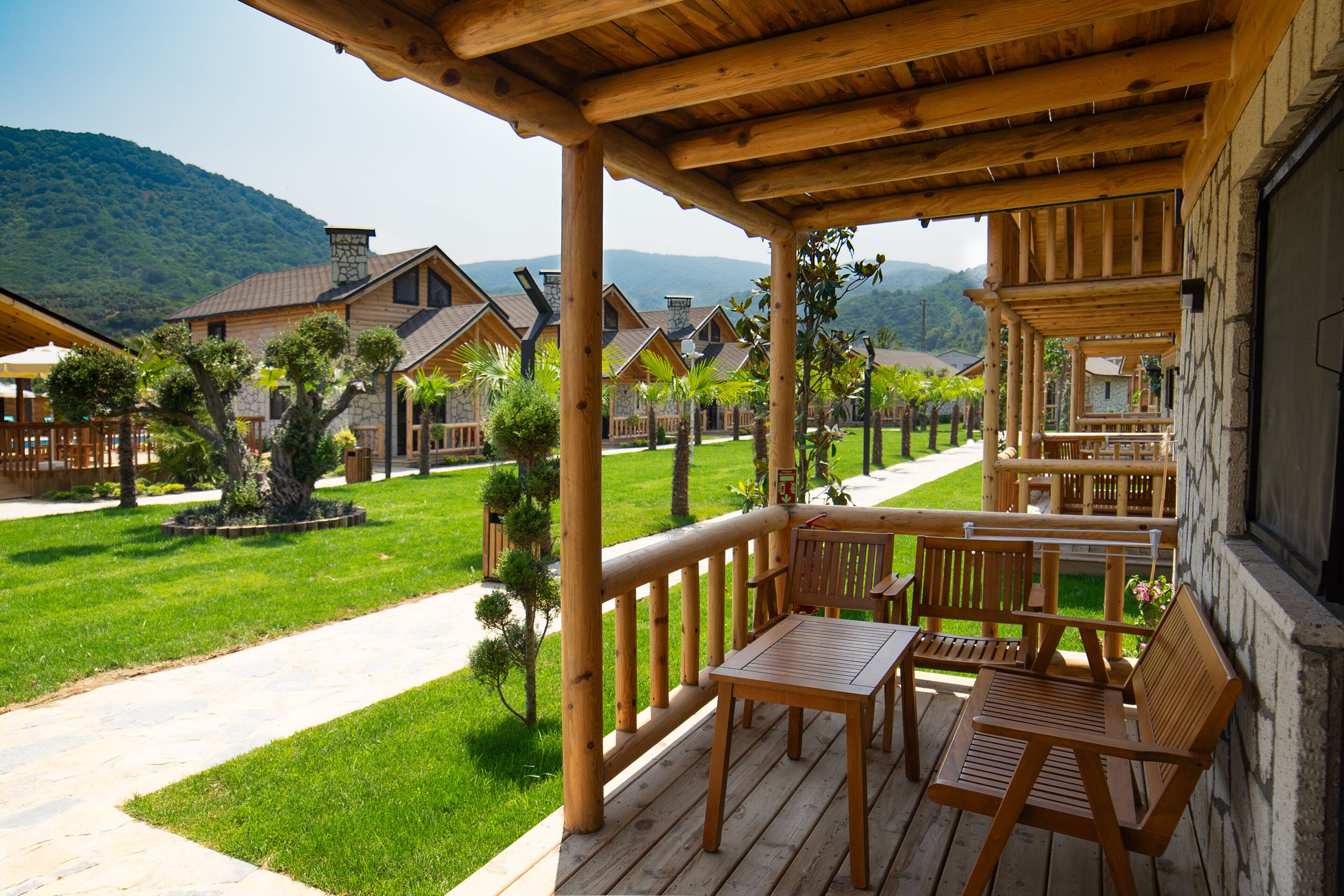 A wooden deck with outdoor seating, surrounded by a well-maintained lawn and trees, overlooking a row of cabins with a mountainous backdrop.