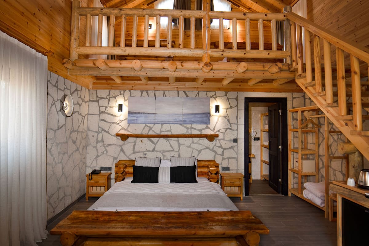 Ein Schlafzimmer in einer Blockhütte mit Steinsicht, einem Holzbett, zwei Nachttischen, einem Beistelltisch, einer Holzbank und einer Treppe.