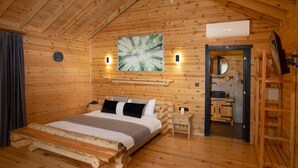 Une chambre à coucher dans une cabane en bois, avec un lit, un tableau, une horloge et une salle de bain visible à travers une porte ouverte.