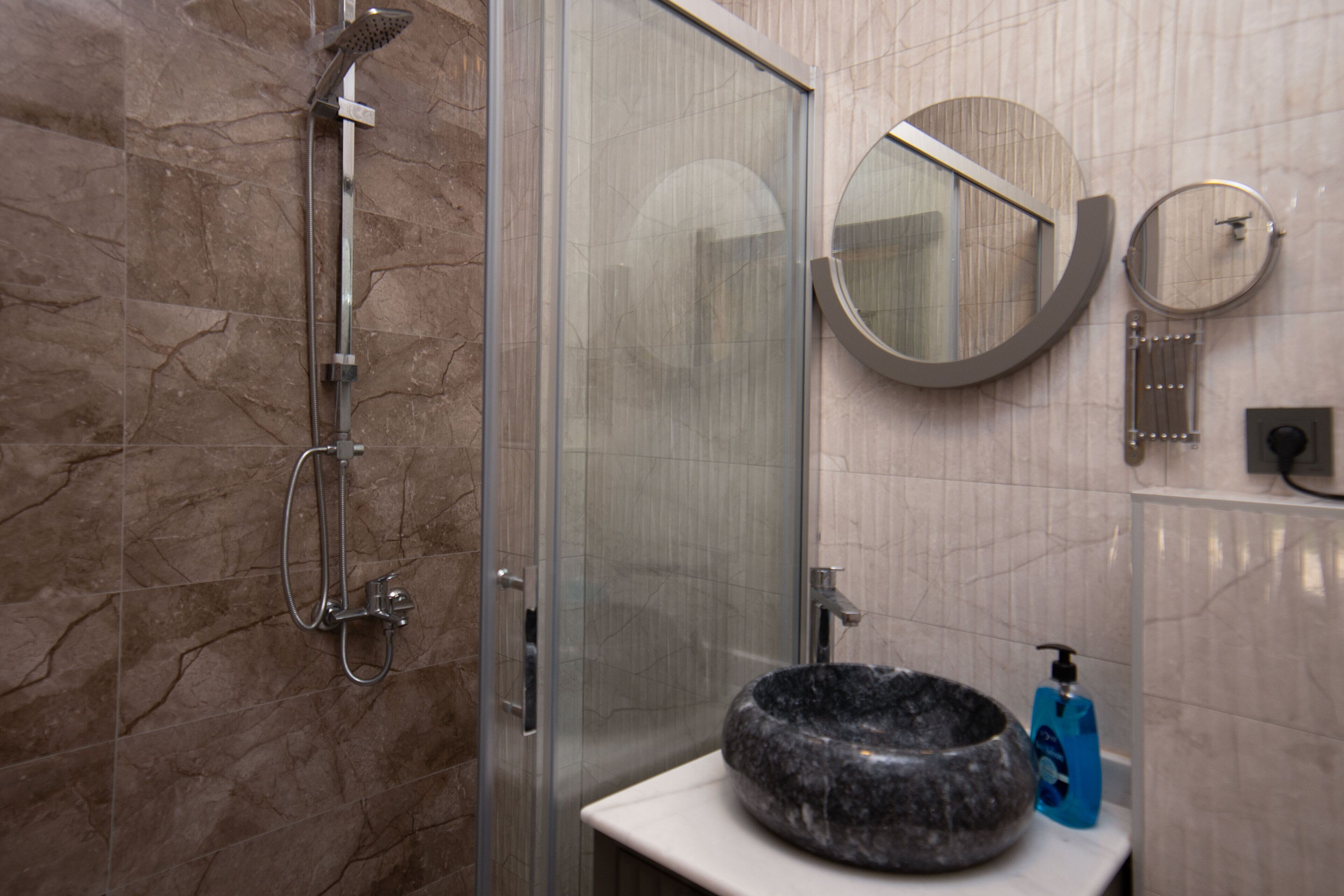 A bathroom with a glass-enclosed shower, a round mirror, a sink with a stone basin, and a blue soap dispenser.