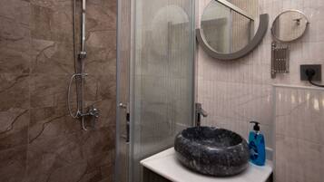 A bathroom with a glass-enclosed shower, a round mirror, a sink with a stone basin, and a blue soap dispenser.