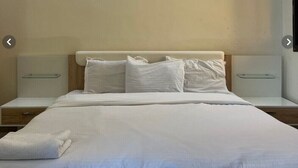 Un llit ben arrenjat amb llençols i almoades blanques, dues taules de nit amb làmpades i una paret senzilla.