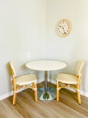 Ein Zimmer mit einem runden, weißen Tisch und zwei Korbstühlen. Eine Holz-Wanduhren ist an der Wand befestigt.