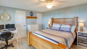 Un dormitorio con una cama de madera, un escritorio con una computadora, una silla, un ventilador de techo y un letrero en la pared.