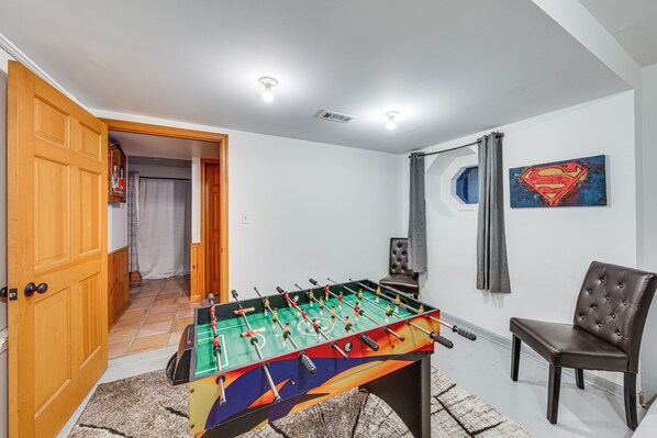 A room with a foosball table, a chair, a Superman painting, and a door leading to another room.
