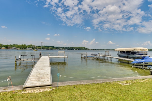 A calm lake with a wooden dock extending into the water, surrounded by grassy areas and a distant tree line.