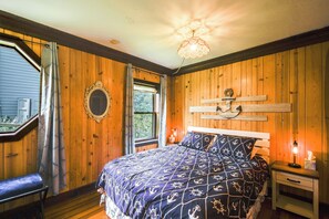 A bedroom with wooden walls, a bed with a nautical-themed bedspread, a bedside table with a lamp, and a window with a view of trees. - Waterfront Fox Lake Vacation Rental w/ Fire Pit! (Fox Lake)