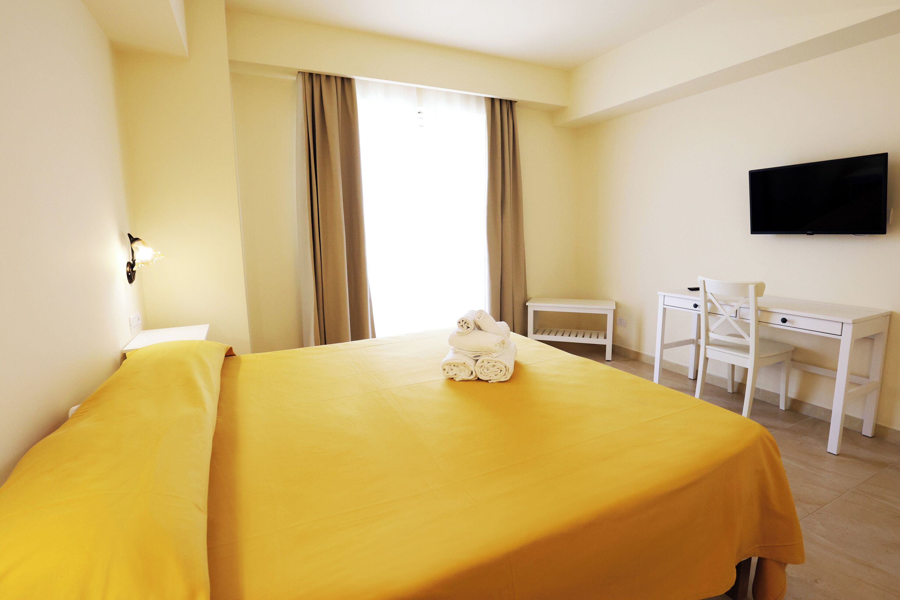 Habitación de hotel con cama, escritorio, silla, televisor y ventana con cortinas.