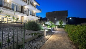 A modern multi-story building with a well-lit exterior, a paved pathway, and landscaped areas with plants and gravel. - Verdello Home Savoca (Savoca)