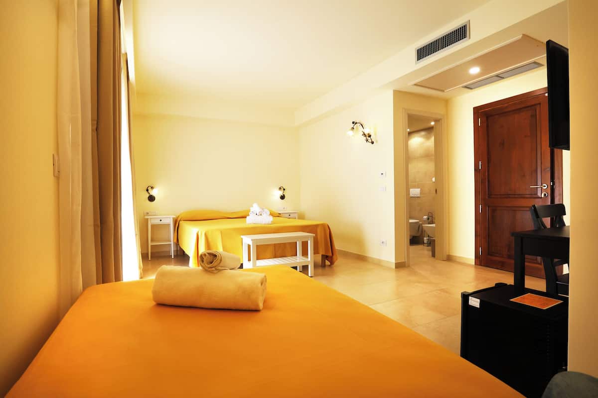 A hotel room with a bed, a small table, a chair, a TV, and a bathroom visible in the background.