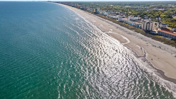 An aerial view of a coastal city with a long beach, buildings along the shore, and a clear blue sky.