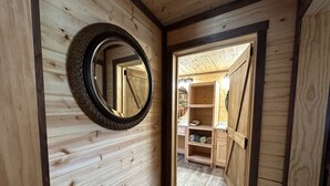 Un interior de madera con un espejo circular en la pared, una puerta de madera que da a una habitación con estanterías y suelo de baldosas.