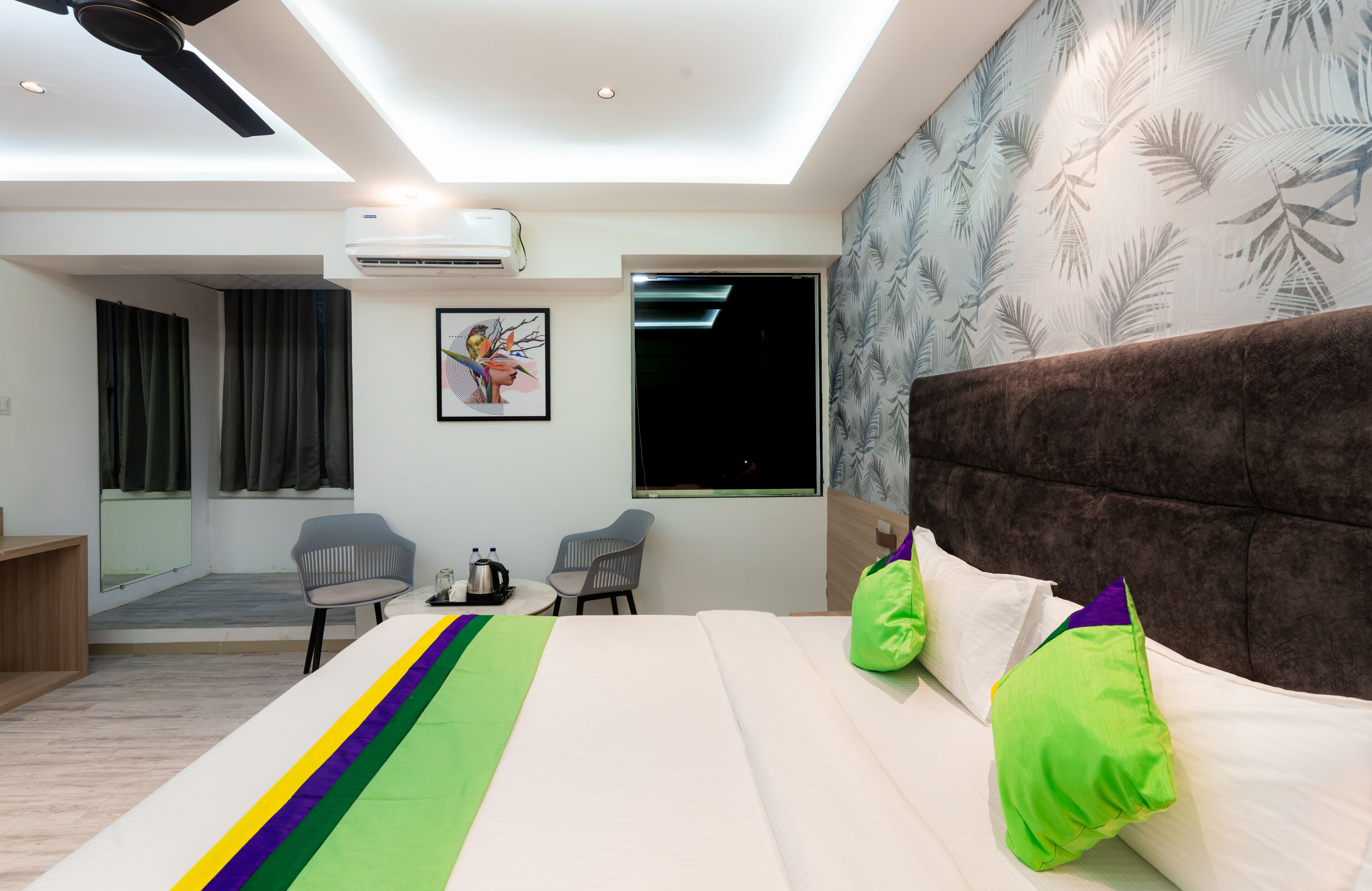 A modern hotel room with a large bed, two chairs, a small table, and a ceiling fan.