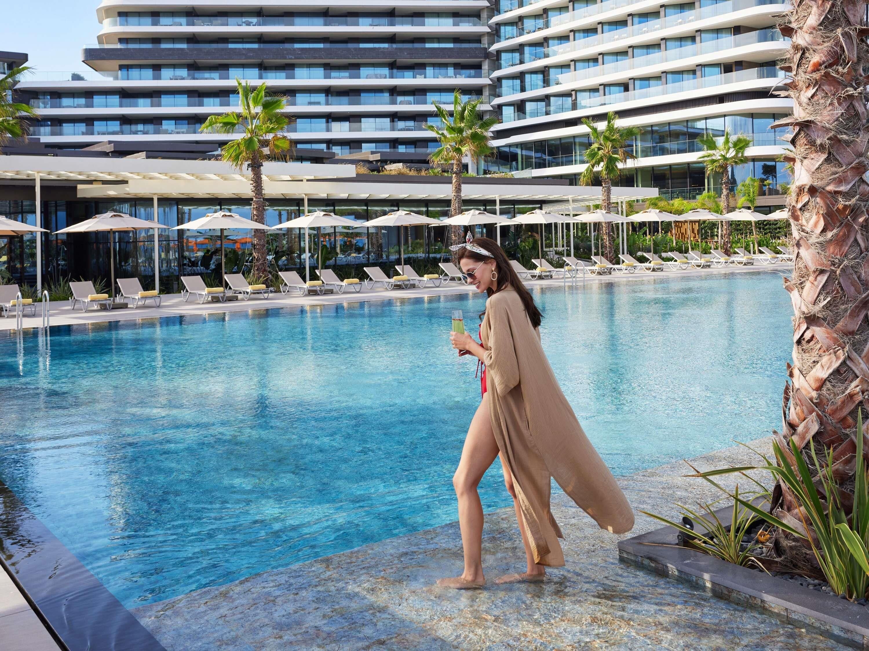 A swimming pool with lounge chairs and umbrellas, surrounded by palm trees and modern buildings.