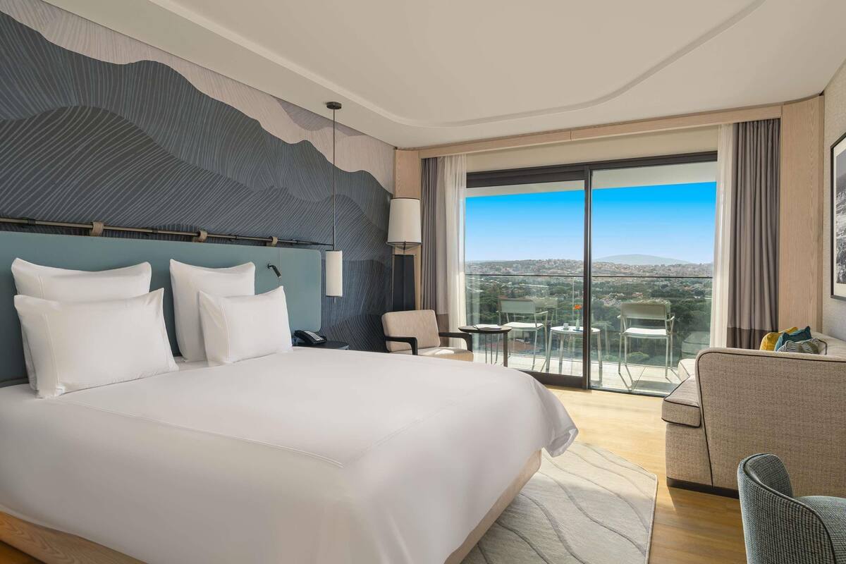 A hotel room with a large bed, a desk, a chair, and a balcony with a city view.