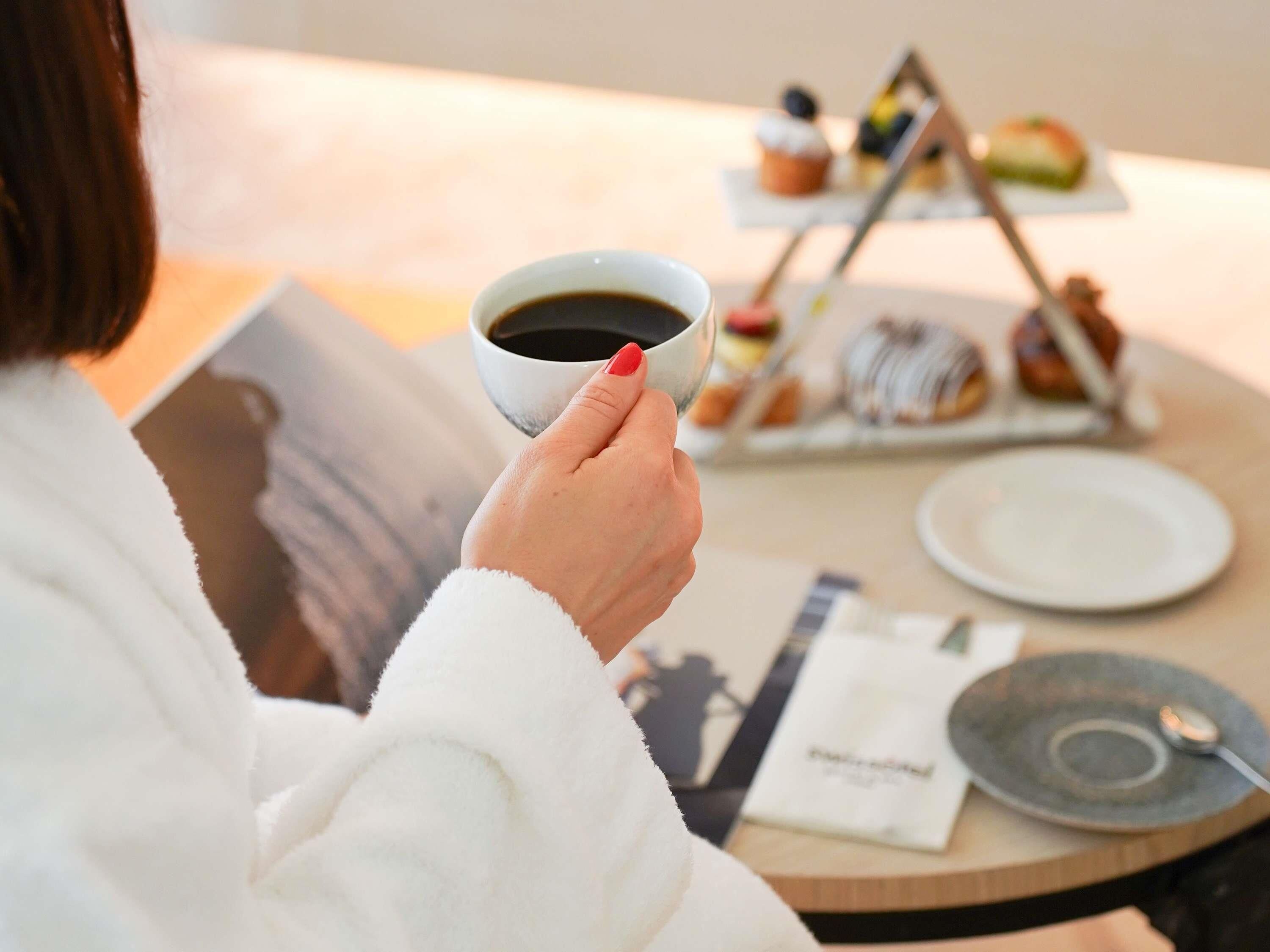 Une personne tenant une tasse de café, une assiette et un plateau de pâtisseries sur une table ronde.