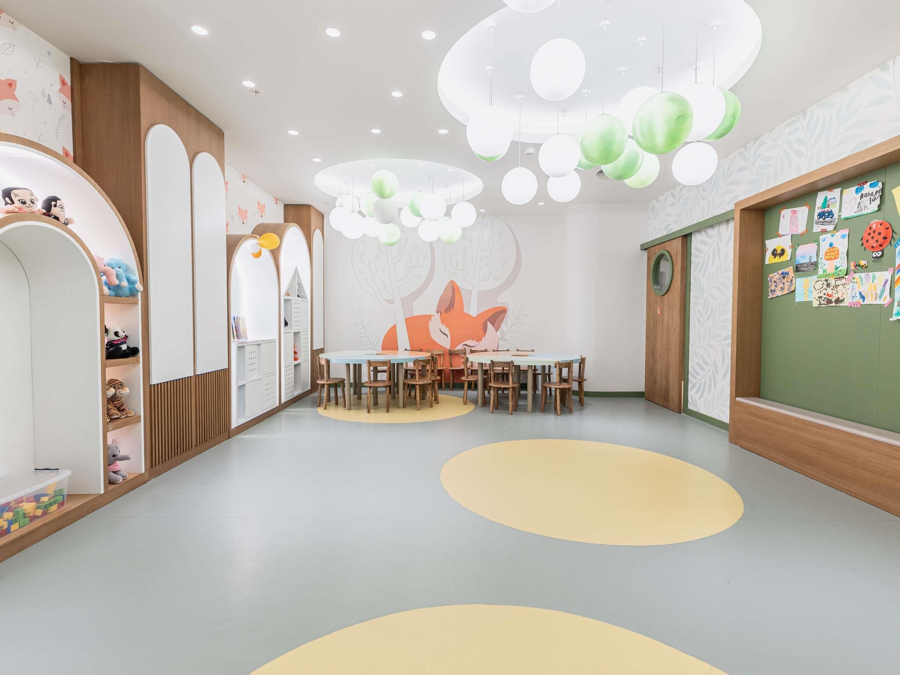 Ein Spielzimmer für Kinder mit einem runden Tisch, Stühlen, Regalen voller Spielzeug und einer Wand mit Kinderzeichnungen.